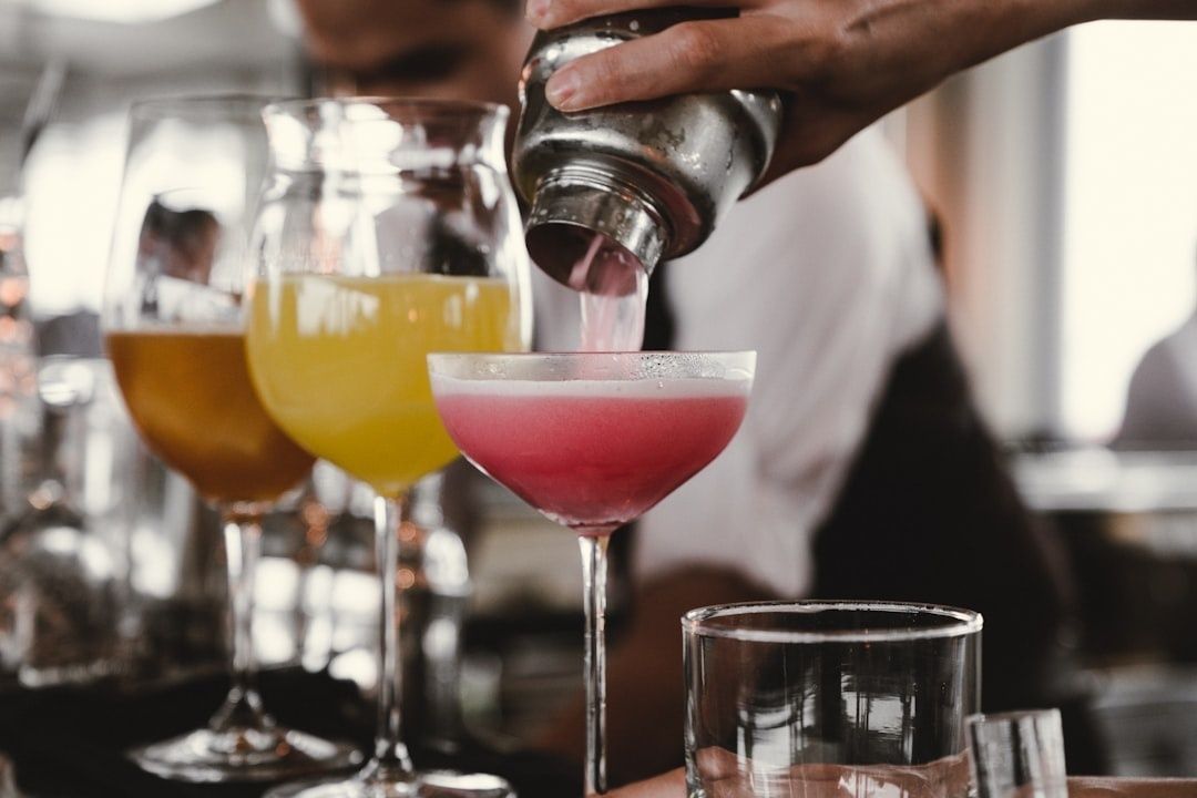 Bartender pours a pink cocktail among colorful drinks in stemmed glasses